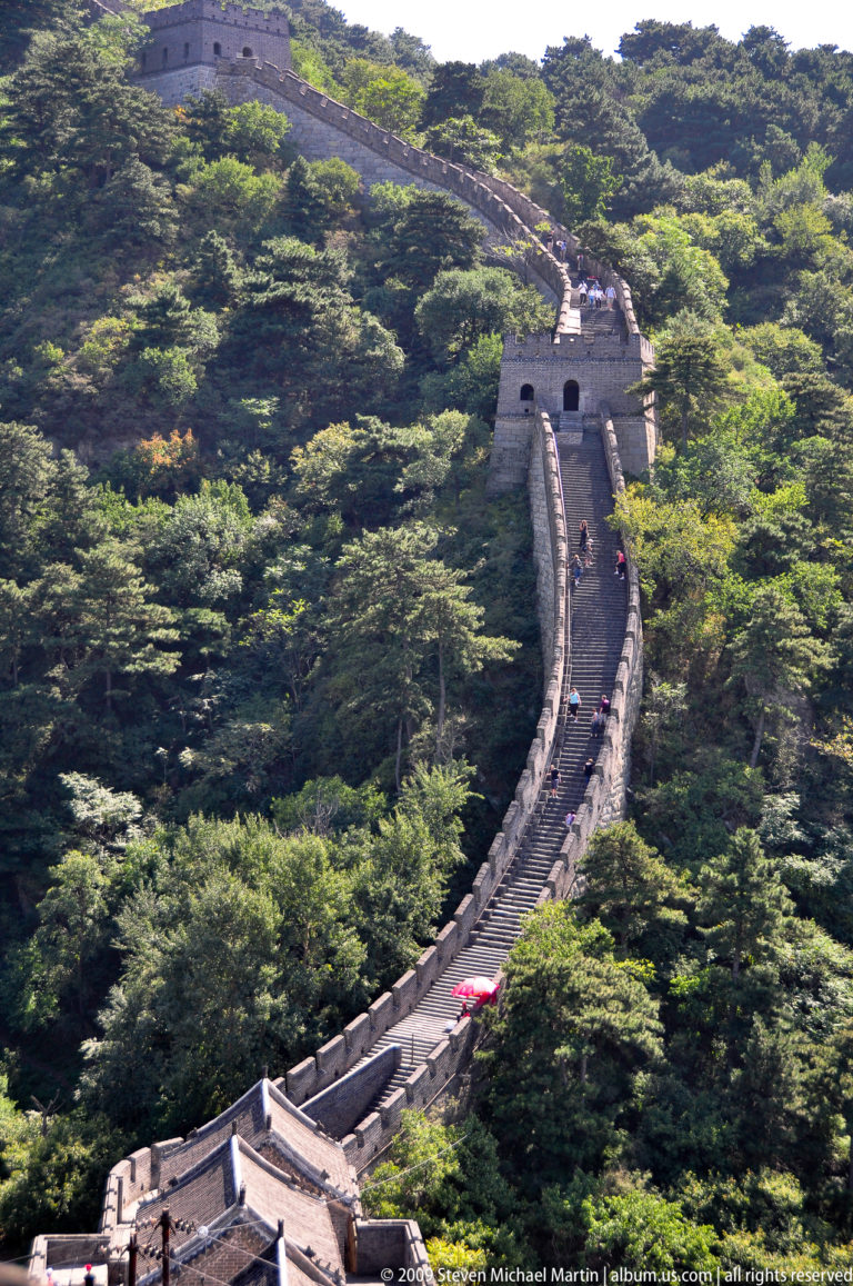 Telephoto view of a very steep Great Wall staircase at Mutianyu rising through forest to a large watchtower, with small visitors spaced along the climb.