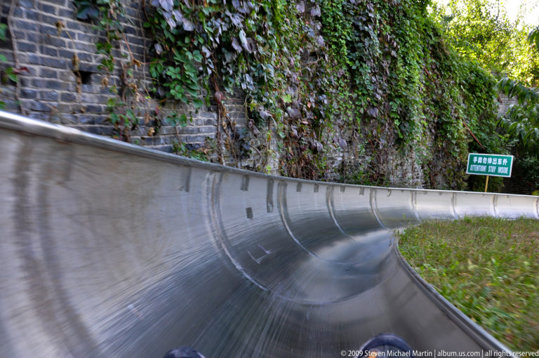 Point-of-view view from a sled descending a curving stainless-steel alpine slide through forest at the Mutianyu Great Wall visitor complex.