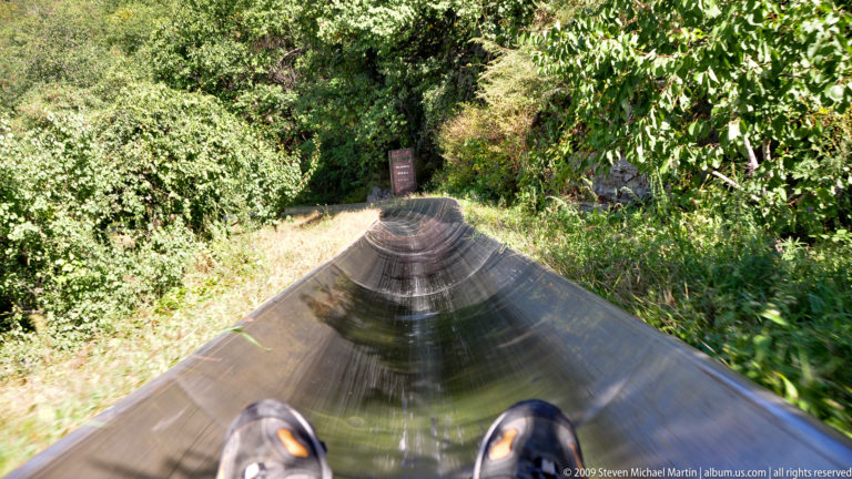 Rider’s point-of-view showing their shoes at the bottom of the frame as a metal alpine slide curves through forest past a warning sign at the Mutianyu Great Wall tourist complex.