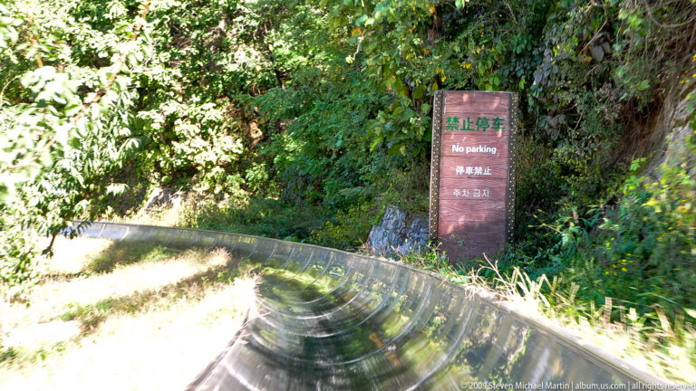 Multilingual brown “No parking” sign beside a curving stainless-steel toboggan track at the Mutianyu Great Wall tourist area.