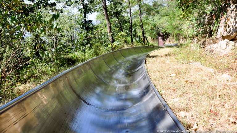 Empty stainless-steel toboggan track curving through dry grass and dense trees on the wooded hillside at Mutianyu.