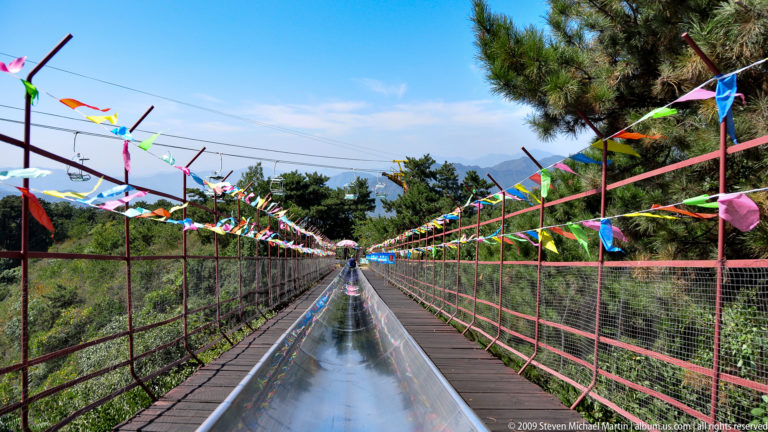 Straight stainless-steel toboggan slide at Mutianyu with parallel wooden walkways, railings, and colorful flags receding in strong central perspective.
