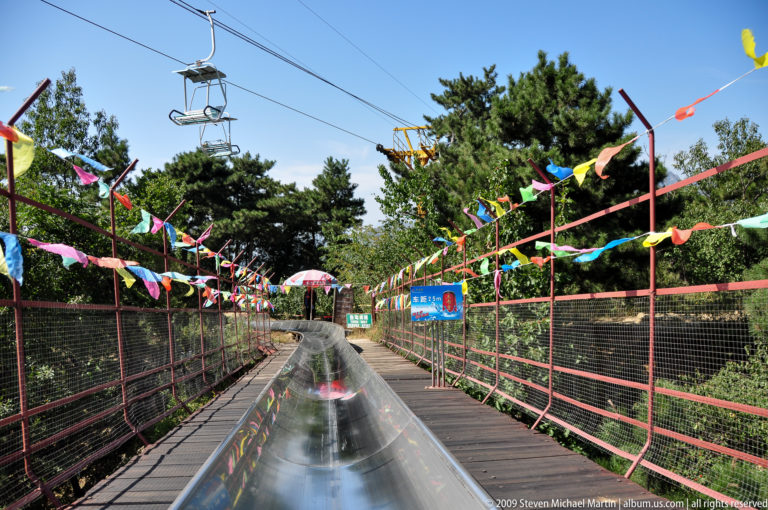Curving stainless-steel alpine slide at Mutianyu passing fencing and colorful flags beneath overhead chairlift cables near a shaded station area.