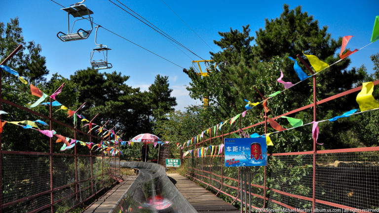 Blue 25 m sign with Chinese text beside a curving stainless-steel toboggan slide, with chairlift chairs passing overhead at Mutianyu.
