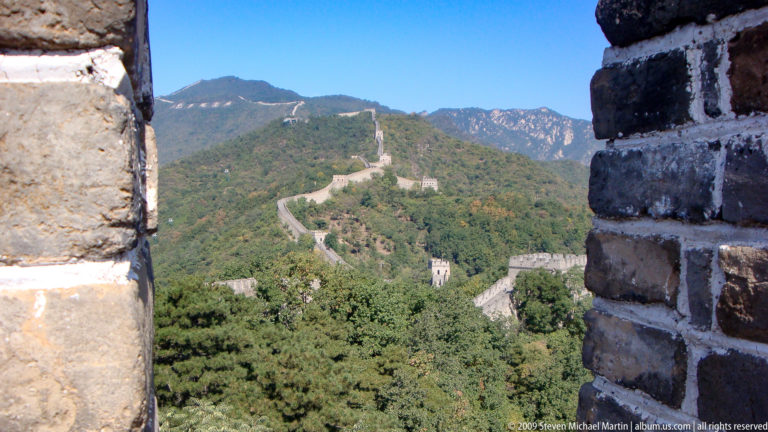 Distant view of the Great Wall at Mutianyu running along green mountain ridges, with multiple square watchtowers under a clear blue sky.