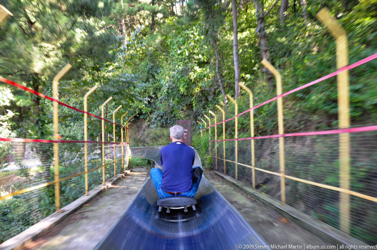 Rear view of a man in a blue shirt riding a small sled down a metal toboggan chute between tall safety fences at the Mutianyu Great Wall area.