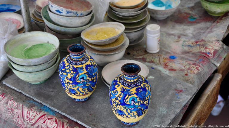 Two small yellow jars with blue dragon designs beside open bowls of colored powders on a stained pottery workshop surface.
