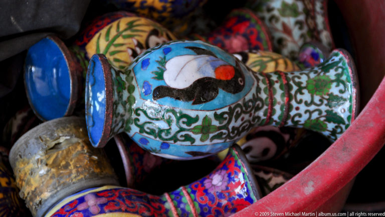 Close-up of a small cloisonne-style vase showing a black-and-white bird among colorful flowers and scrolling leaves on a blue background.
