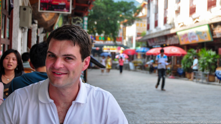 Smiling young male visitor in a white shirt seated on Yangshuo’s West Street with cafés and umbrellas in the busy pedestrian street behind him.