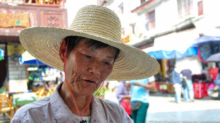 Close candid portrait of an older woman in a wide woven straw hat looking downward, with colorful market stalls and pedestrians blurred behind her in Guilin.