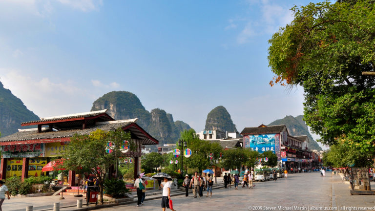 Pedestrian plaza in Yangshuo with low-rise shops in traditional-style roofs and karst hills rising behind the town.