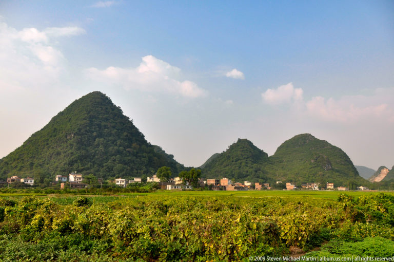 Rural farming village at the base of rounded limestone hills, with green cultivated fields in the foreground.