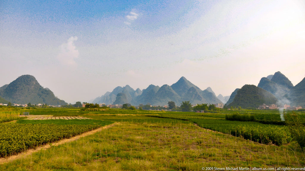 Green and gold cultivated fields with parallel boundaries leading toward hazy, layered karst peaks near Guilin, China.