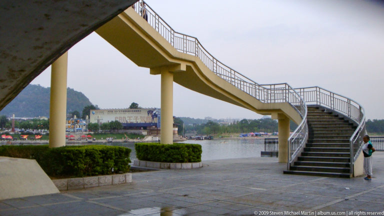 Curving elevated stair bridge beside a lake in Guilin with distant karst hills in the background.