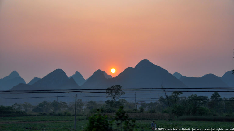 Hazy orange sun setting behind dark karst hills with power lines crossing above farm fields near Guilin.