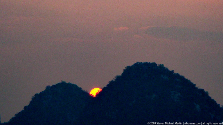 Close telephoto view of an orange sun partly hidden behind a dark wooded karst ridge in a hazy Guilin evening sky.