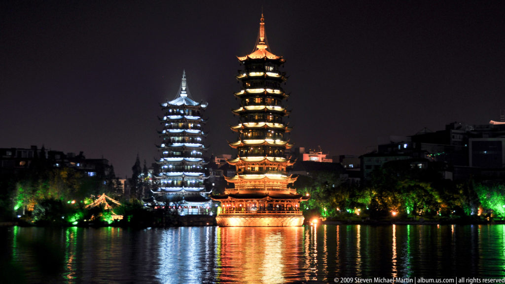 Night view of the illuminated Sun and Moon Pagodas on Shan Lake in Guilin, one glowing gold and the other white, reflected in the dark water.