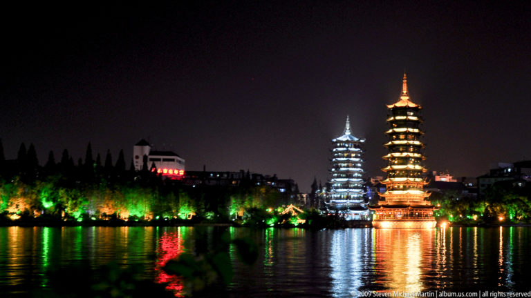 Night view of the illuminated Sun and Moon Pagodas on Shan Lake in Guilin, one glowing gold and the other white-blue with reflections in the dark water.