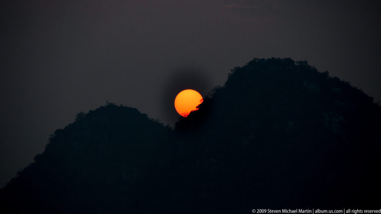 Deep orange sun more than half visible as it sets behind the dark wooded ridge of a Guilin karst hill against a gray-orange sky.
