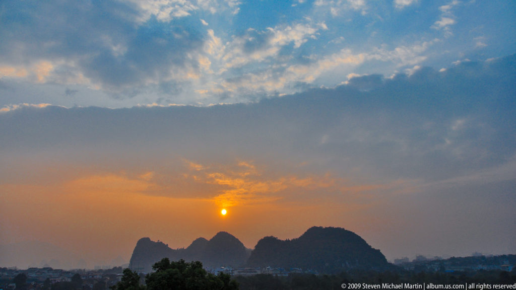 Orange sun setting above hazy limestone hills with low urban development at the edge of Guilin.