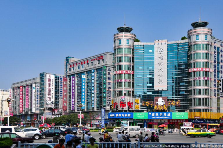 Glass-fronted shopping towers above a busy intersection in downtown Guilin with cars, buses, bicycles, and vertical Chinese signs on the facades.