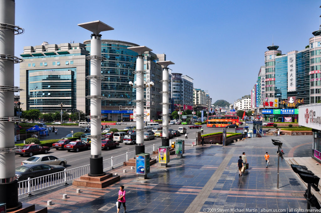 Wide paved plaza in Guilin with tall cylindrical light columns beside a multi-lane road and modern glass commercial buildings.