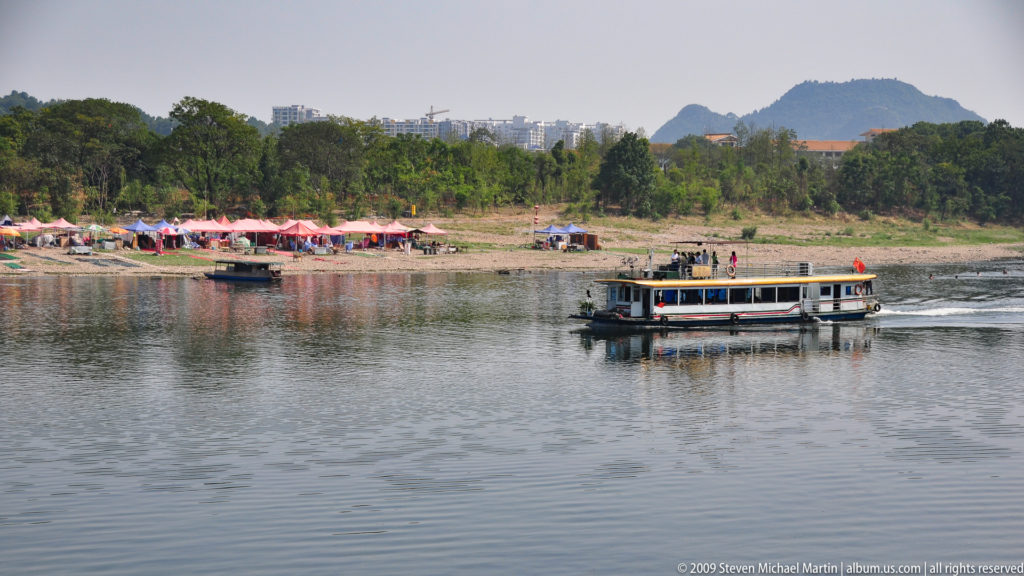 Passenger tour boat with Chinese flags crossing calm water in Guilin, with karst hills in the background.