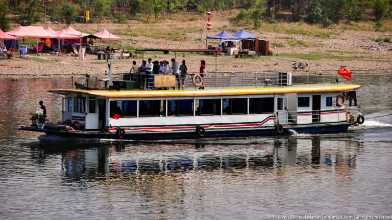 Passenger tour boat with enclosed cabin and open upper deck crosses calm water near a developed shoreline with karst hills in the distance in Guilin, China.