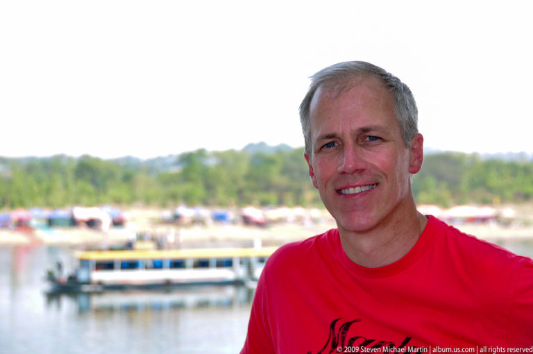Smiling gray-haired man in a red shirt near the Guilin waterfront with a blurred tour boat and distant tree-lined shore in the background.