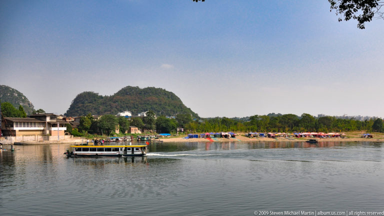 Passenger tour boat with enclosed cabin and open upper deck crossing calm water near a developed shoreline and distant karst hills in Guilin.
