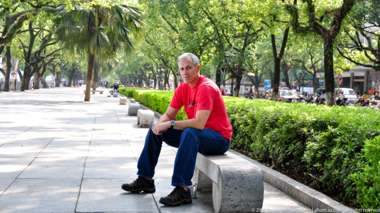 Gray-haired man in a red shirt sitting beside a clipped hedge on a tree-lined promenade in a Guilin park.