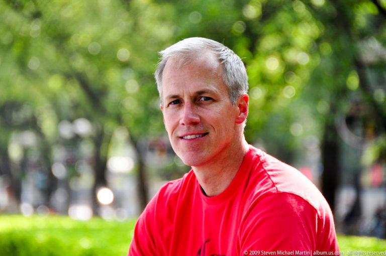 Gray-haired visitor in a red shirt sitting by a clipped hedge on a tree-lined promenade in a Guilin park.