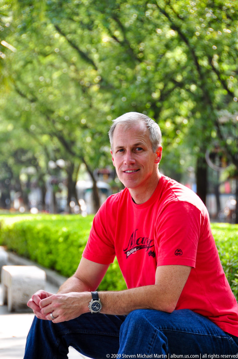 Gray-haired man in a red shirt sitting beside a hedge on a tree-lined promenade in a Guilin park.