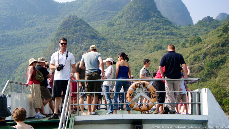 Tourists stand along the railings of an open upper deck on a Li River cruise boat with steep karst mountains in the background.