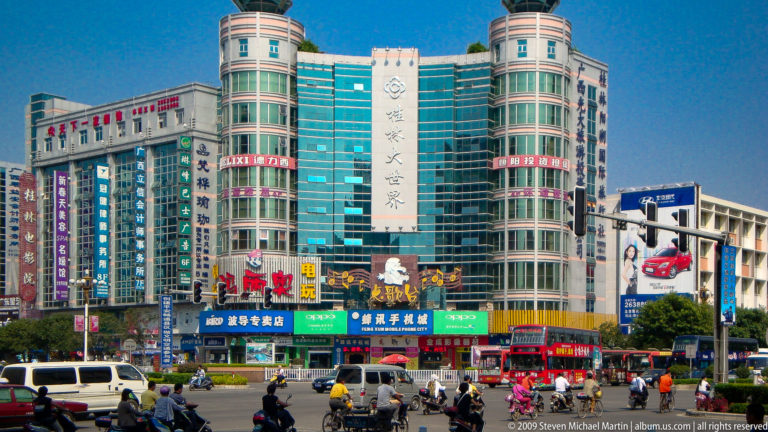 Turquoise-glass shopping complex at a busy intersection with buses and bicycles in downtown Guilin, China.