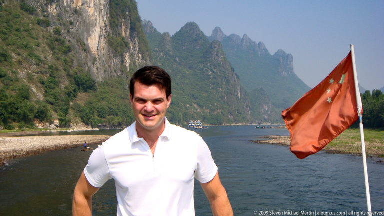 Man in a white shirt standing on a Li River cruise boat with steep karst peaks rising along both banks.