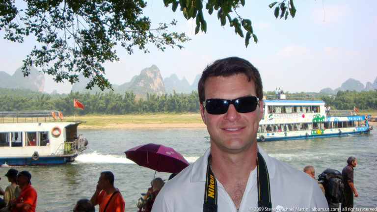 Male traveler in dark sunglasses with a camera strap stands under tree branches on a Li River cruise boat, with a multi-deck passenger boat and karst mountains in the background.