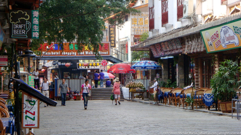Stone-paved pedestrian street in Yangshuo lined with cafés, umbrellas, and low-rise traditional-style buildings with English and Chinese shop signs.