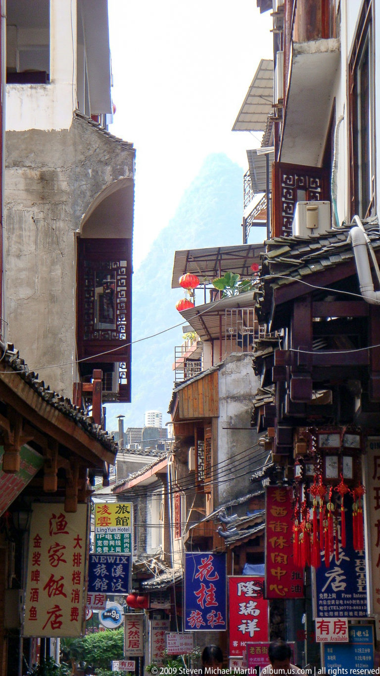 Wooden balconies with red lanterns and hanging signs on a narrow commercial street in Yangshuo, with karst cliffs faintly visible between the buildings.