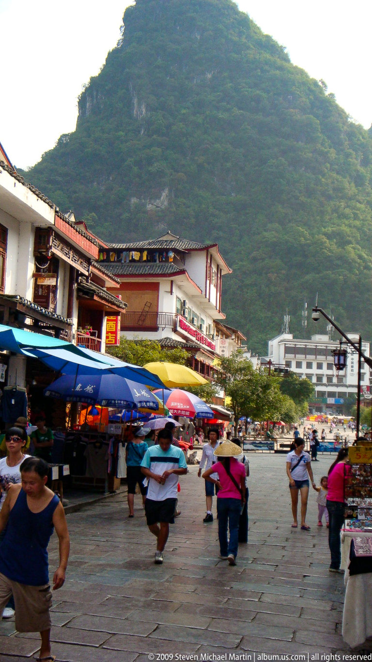 Busy Yangshuo shopping street with colorful umbrellas and low-rise buildings beneath a steep green karst hill.