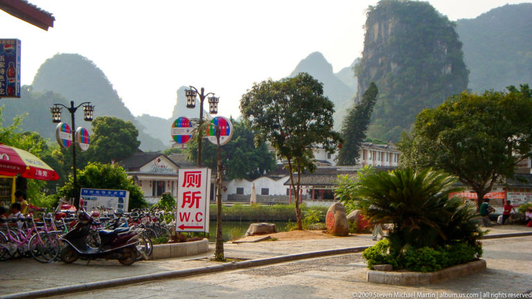 Bilingual W.C. toilet sign near parked scooters with karst cliffs and low buildings in a Yangshuo town.