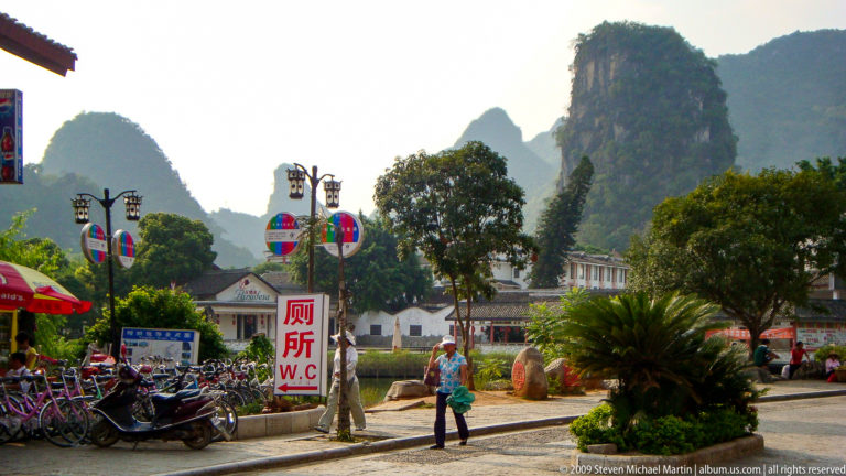 Bilingual toilet sign reading 厕所 and W.C. beside parked scooters in a Guilin-area town with a steep karst hill rising behind low buildings.