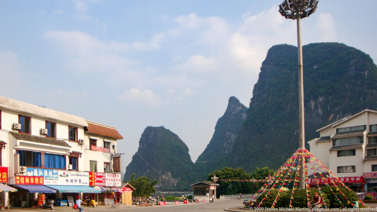 Urban plaza in Yangshuo with colorful conical streamer display and road leading toward steep karst peaks.