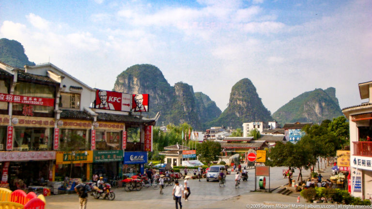 Busy commercial street in Yangshuo with a prominent KFC sign, local shops, scooters, and pedestrians beneath steep karst peaks.