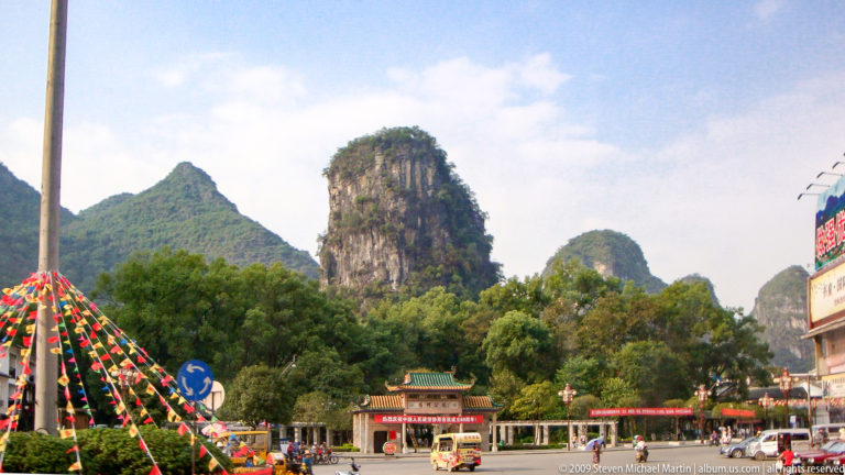 Steep vegetated limestone tower rising behind a decorated roundabout at the edge of a town.