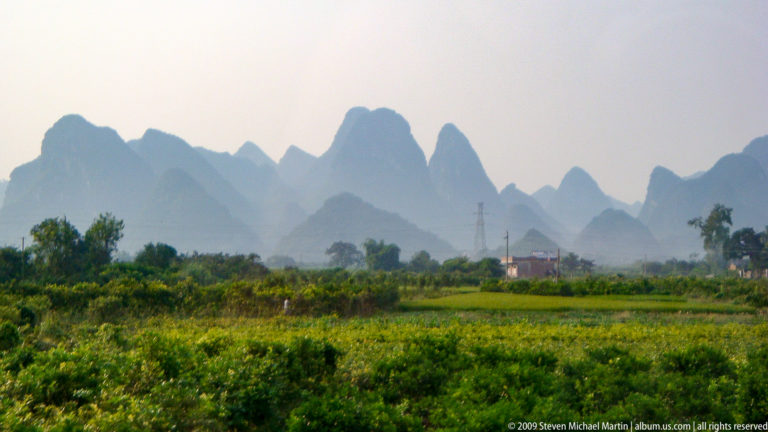 Green crop fields on an alluvial plain leading toward layered blue-gray karst peaks in hazy light, with scattered buildings and utility poles in the distance.