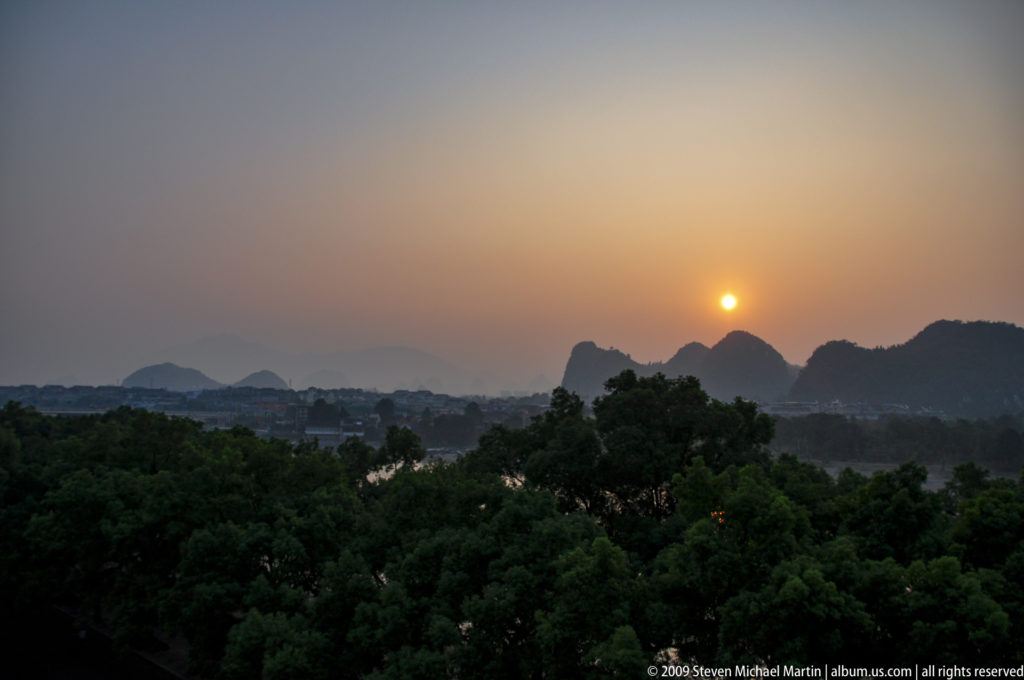 Orange sun setting behind hazy, silhouetted limestone peaks in Guilin with dark trees in the foreground.