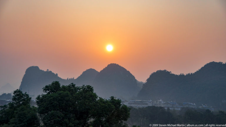 Orange sun setting through haze above silhouetted limestone peaks and trees in Guilin.