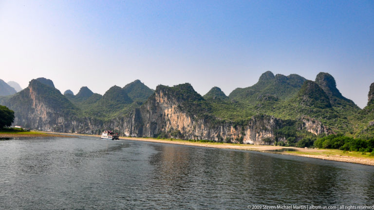 Wide bend of the Li River flowing past pale limestone cliffs and forested karst hills with exposed gravel bars along the banks.