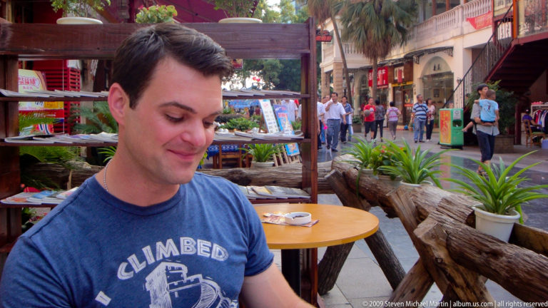 Young male traveler in a blue T-shirt seated at a round wooden table at an outdoor café on a busy pedestrian street in Guilin.